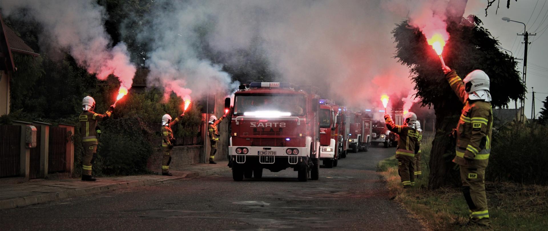 Pojazdy pożarnicze jadą jeden z drugim. Po bokach drogi stoją strażacy z odpalonymi racami