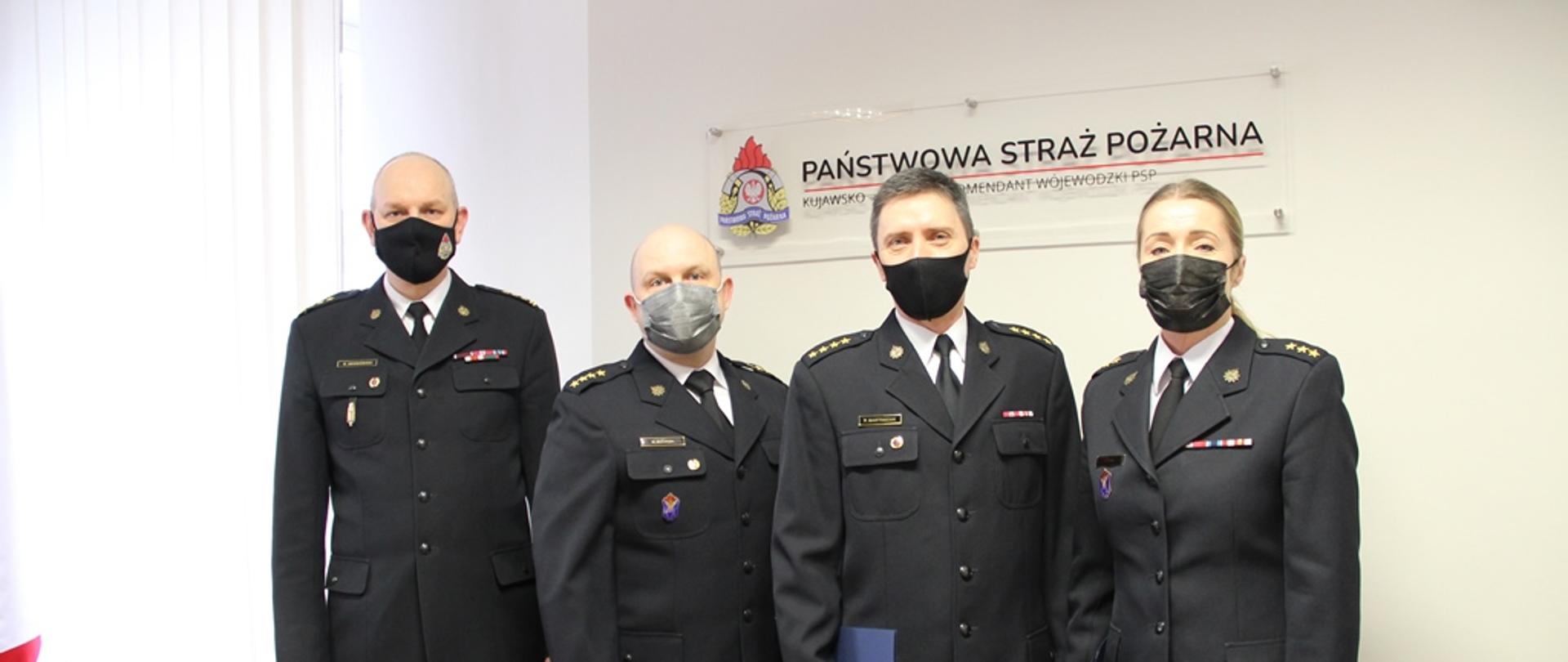 Na zdjęciu trzech funkcjonariuszy PSP, którzy odebrali nagrody jubileuszowe, w towarzystwie zastępcy komendanta wojewódzkiego. Tłem jest logo PSP.