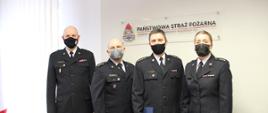 Na zdjęciu trzech funkcjonariuszy PSP, którzy odebrali nagrody jubileuszowe, w towarzystwie zastępcy komendanta wojewódzkiego. Tłem jest logo PSP.