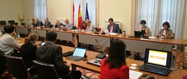 Konsultacje Pełnomocników ds. otwartości danych z instytucjami hiszpańskimi. Ludzie siedzą przy owalnym stole. W tle flagi Polski, UE i Hiszpanii.