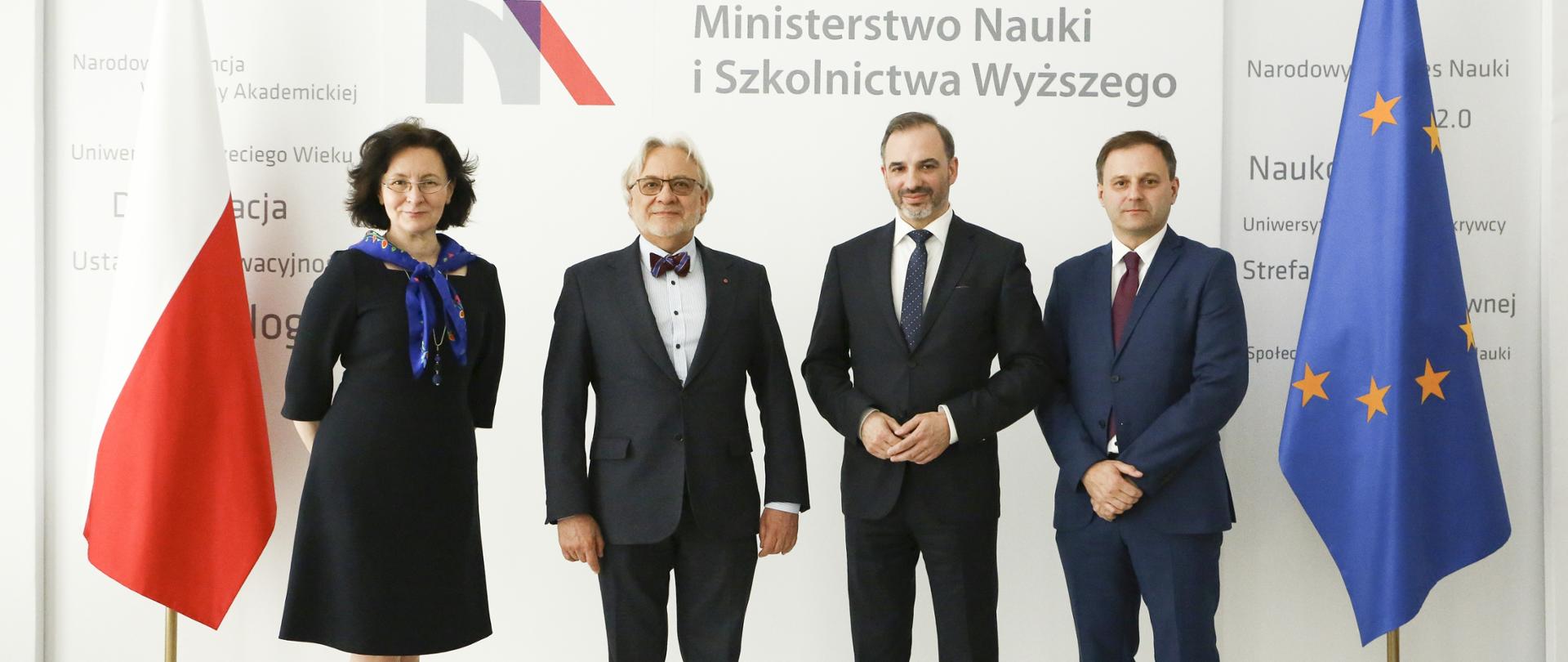 Uczestnicy spotkania stoją na tle ściany, za nimi flagi Polski i UE