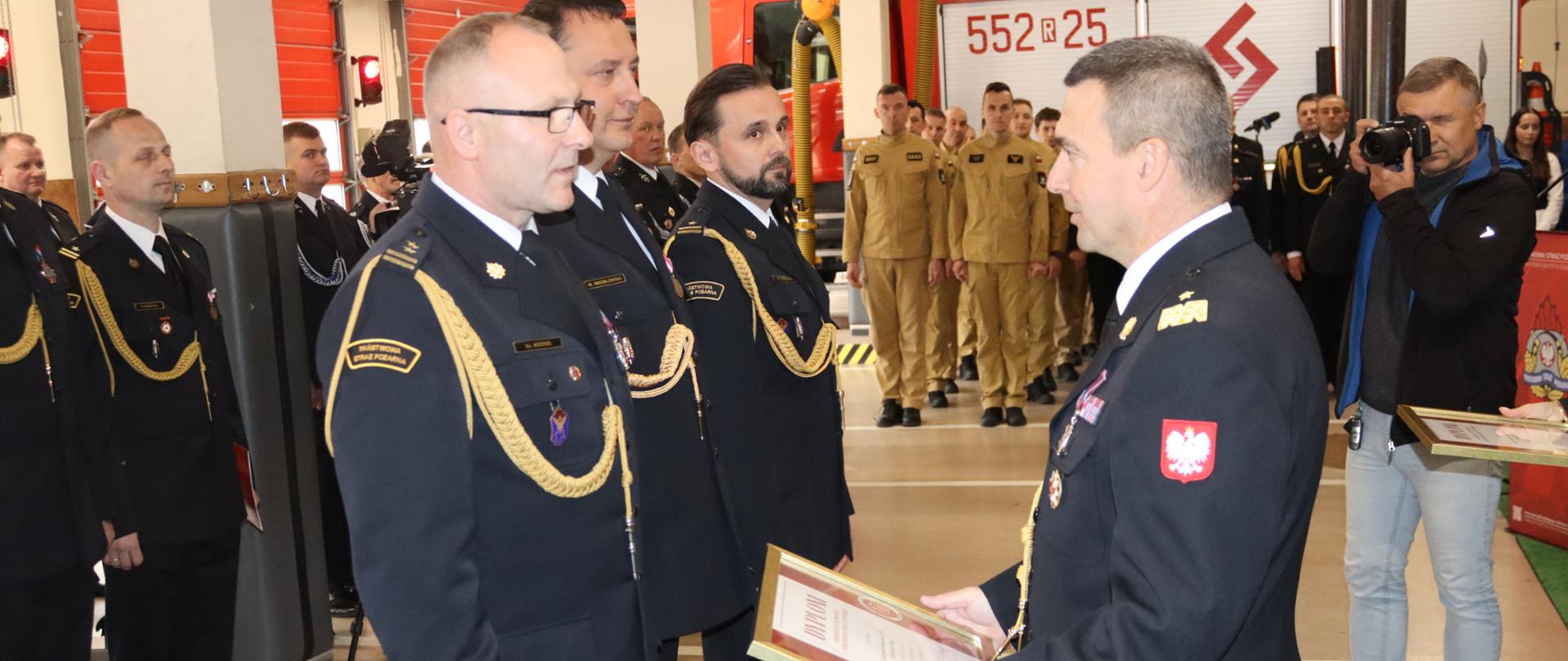 Zdjęcie zrobione w jednostce ratowniczo-gaśniczej podczas uroczystej zbiórki okazji Dnia Strażaka. Na zdjęciu strażacy w galowych mundurach oraz osoby cywilne uczestniczące w uroczystości.