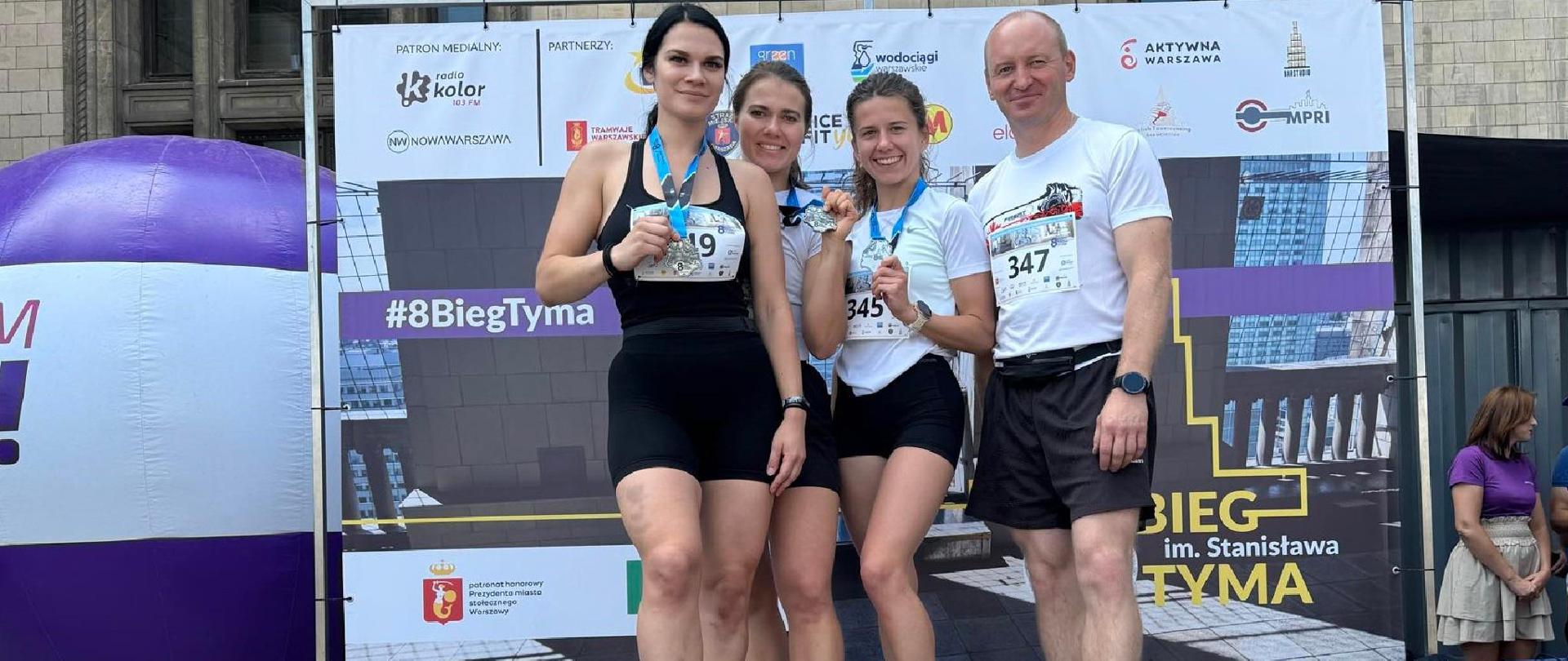 Zdjęcie pamiątkowe uczestników biegu. Na ustawionym przed Pałacem Kultury i Nauki w Warszawie podium stoją 4 osoby (trzy kobiety i mężczyzna) – członkowie reprezentacji SA PSP w Krakowie. Kobiety mają na szyjach pamiątkowe medale.