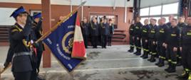 Zdjęcie przedstawia zbiórkę strażaków w pomieszczeniu garażowym. Po prawej stronie w dwuszeregu stoją strażacy w czarnych mundurach koszarowych, na wprost również w dwuszeregu strażacy w czarnych mundurach galowych. Po lewej poczet ze sztandarem.