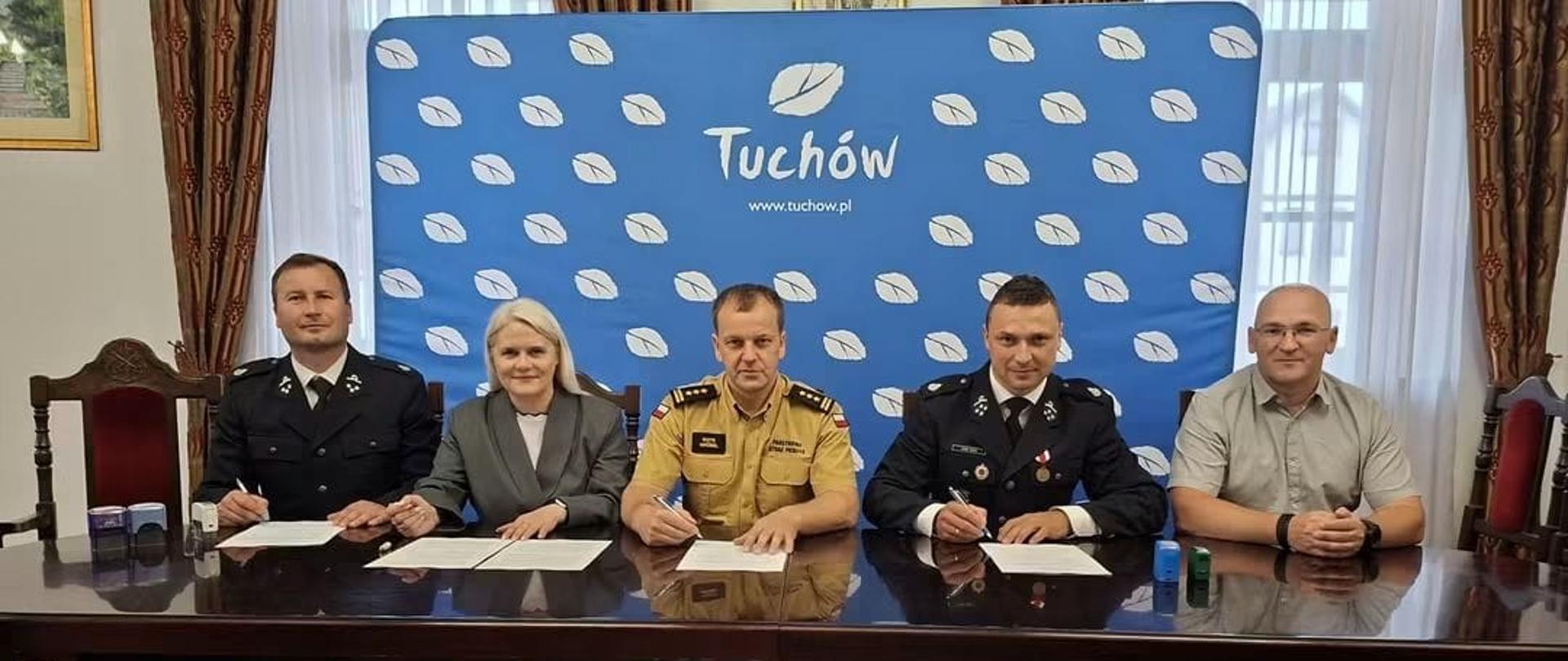 Uroczyste podpisanie porozumienia włączenia w struktury KSRG Jednostki OSP Piotrkowice w Urzędzie Gminy Tuchów. Od lewej siedzą: przedstawiciel OSP Piotrkowice – Naczelnik, Burmistrz Tuchowa, Zastępca Komendanta Miejskiego PSP w Tarnowie, drugi przedstawiciel OSP Piotrkowice – prezes oraz Komendant Gminny OSP. W tle niebieska ścianka promocyjna z logo Tuchowa.