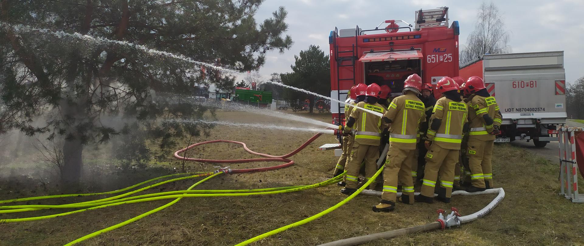 Zdjęcie przedstawia strażaków stojących przy samochodzie strażackim podczas ćwiczeń. Strażacy podają wodę przez prądownice. Na zdjęciu widać kilka linii wężowych. 