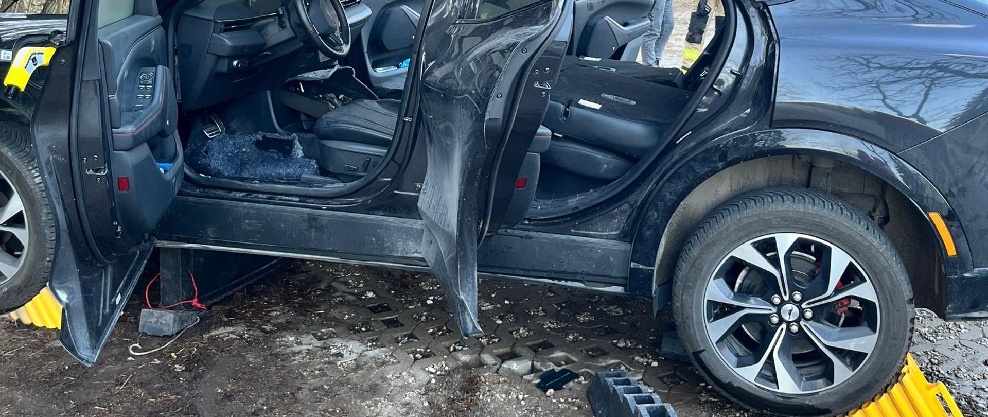 Zdjęcie przedstawia samochód osobowy w którym doszło do pożaru wnętrza. Auto ma unieruchomione koła żółtymi klinami oraz stoi na klinach schodkowych znajdujących się pod pojazdem.