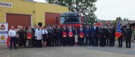 Zdjęcie przedstawia wszystkie zgromadzone osoby podczas przekazania nowego samochodu ratowniczo-gaśniczego. Zdjęcie wykonane zostało na tle remizy strażackiej oraz nowego samochodu.