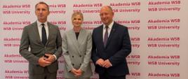 Dwaj mężczyźni i stojąca pomiędzy nimi kobieta pozują do zdjęcia na tle białej ścianki z logo Akademii WSB.