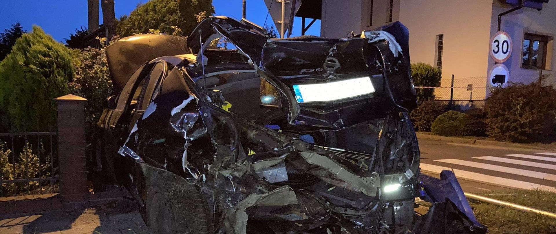 Zdjęcie przedstawia rozbity pojazd marki Nissan Primera koloru ciemnego. Pojazd z rozbitym tyłem stoi na pasie zieleni prostopadle do jezdni, a przód pojazdu znajduje się na posesji prywatnej. Pojazd wjeżdżając na posesję uszkodził ogrodzenie.