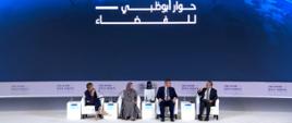 Dwie kobiety, jedna w szarym hidżabie, druga w granatowym ubraniu, i dwóch mężczyzn w garniturach siedzi na białych fotelach w rzędzie i rozmawia, za nimi na ścianie wielki ekran z napisem Abu Dhabi - space debate.