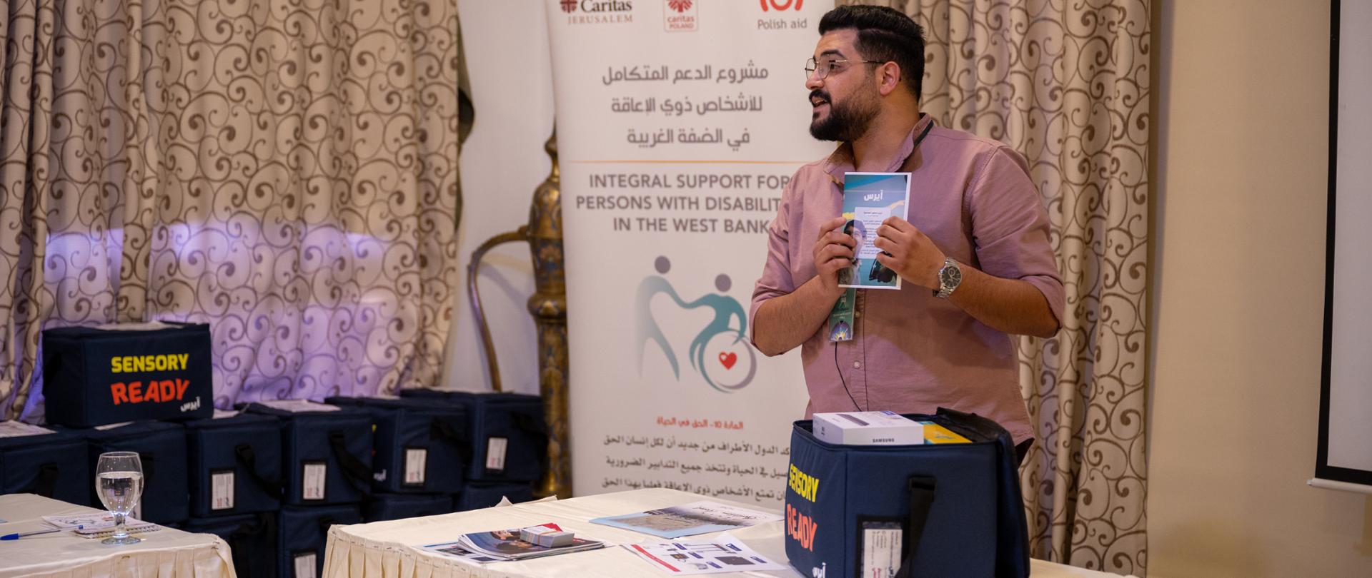 Obok stołu stoi mężczyzna i prezentuje zawartość torby z zestawem sensorycznym