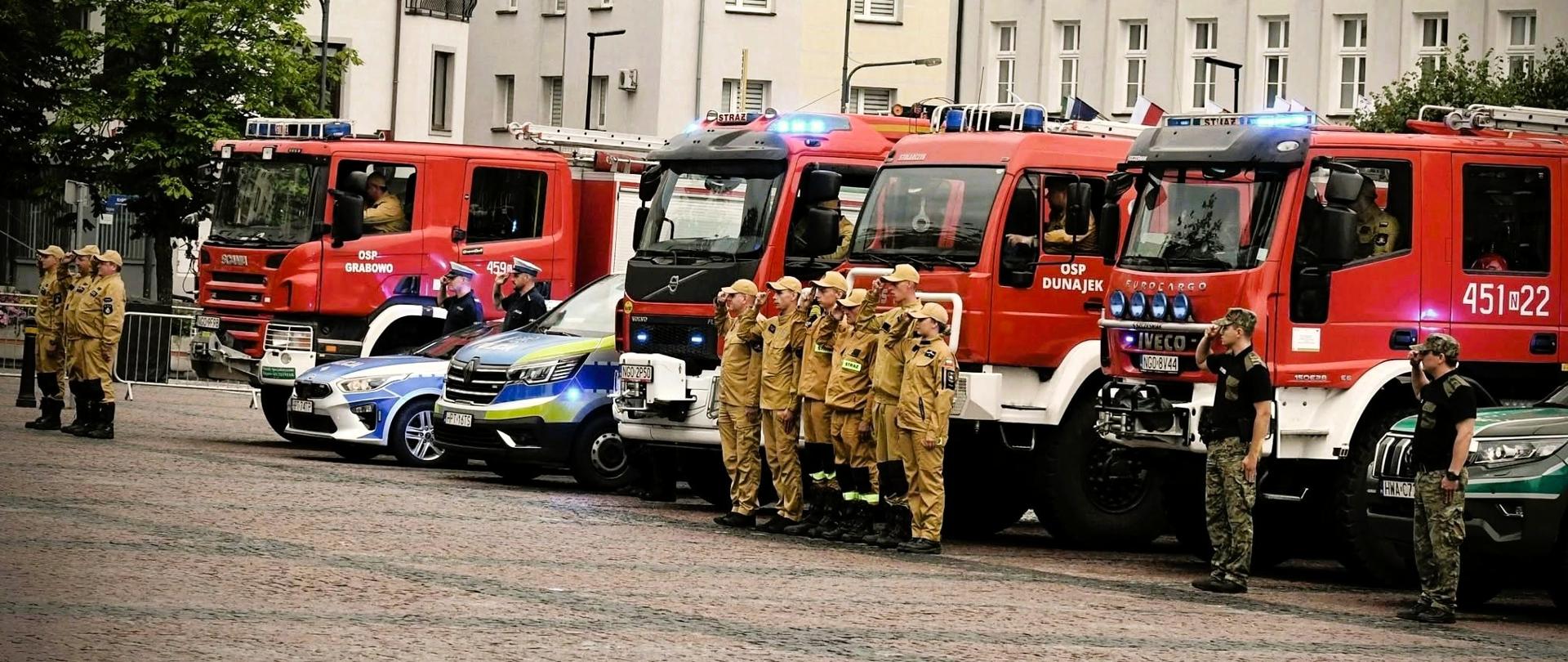 Zdjęcie przedstawia strażaków z Komendy Powiatowej Państwowej Straży Pożarnej w Gołdapi oraz druhów jednostek Ochotniczych Straży Pożarnych gminy Gołdap uczestniczących w obchodach organizowanych na Placu Zwycięstwa w Gołdapi upamiętniających 82. rocznicę wybuchu Powstania Warszawskiego uruchamiając o godz. 17:00 (godzinie „W”) sygnały świetlne i dźwiękowe w pojazdach pożarniczych