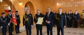 Prezes jednostki OSP Folwarki prezentuje akt włączenia jednostki do KSRG oprawiony w ramce. Na planie zdjęciowym 4 osoby. 