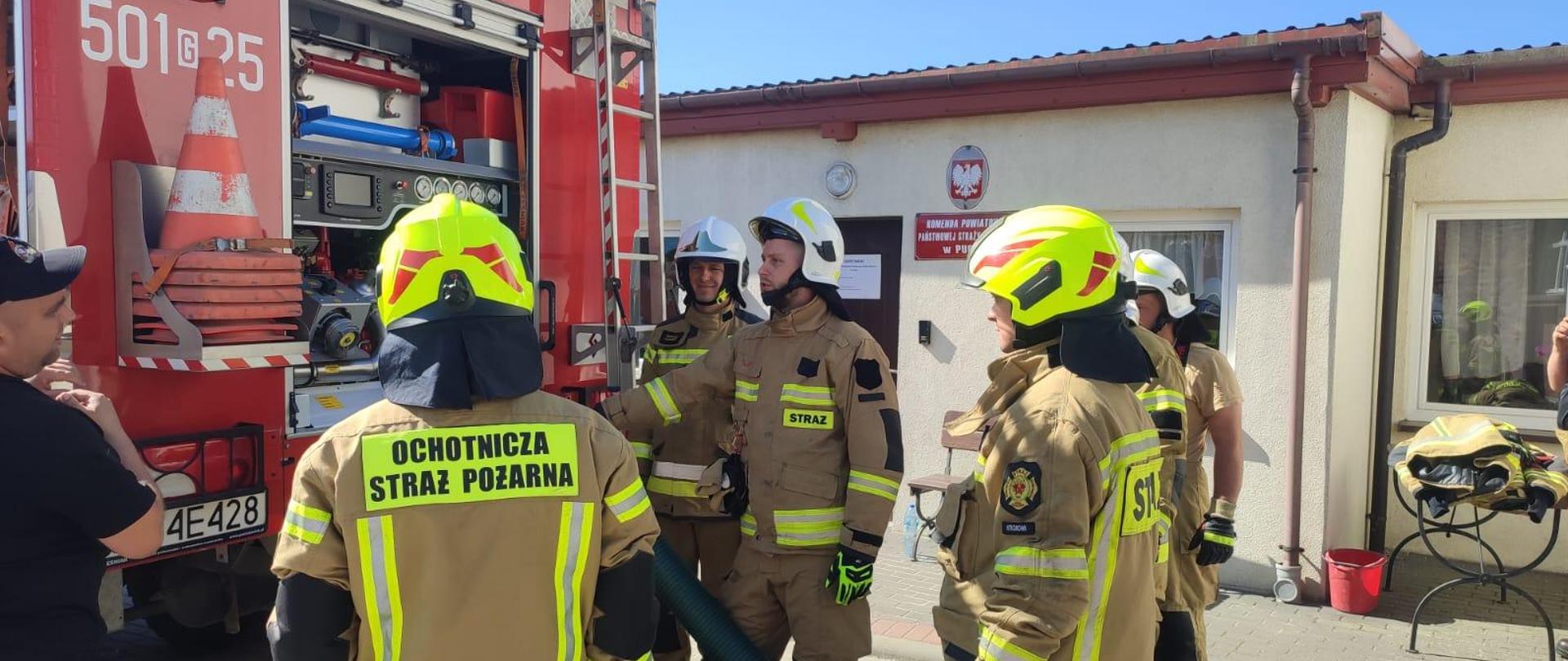 Na zdjęciu druhowie OSP ubrani w ubranie bojowe z helmami na głowie stoją za wozem bojowym i obserwują autopompę podczas pracy. 