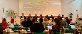 Jest to ujęcie z perspektywy uczestnika siedzącego w sali konferencyjnej, skierowane na stół prezydialny i główny ekran. Na ekranie widnieje napis: „Convention on Wetlands, Ramsar 67th meeting of the Standing Committee, 4–8 May 2026”, z grafiką mokradeł w tle. Przy stole na podwyższeniu siedzi osiem osób prowadzących spotkanie. Przed nimi, w dolnej części kadru, widać głowy i ramiona słuchaczy oraz ich stanowiska pracy z rozstawionymi laptopami i notatkami. 