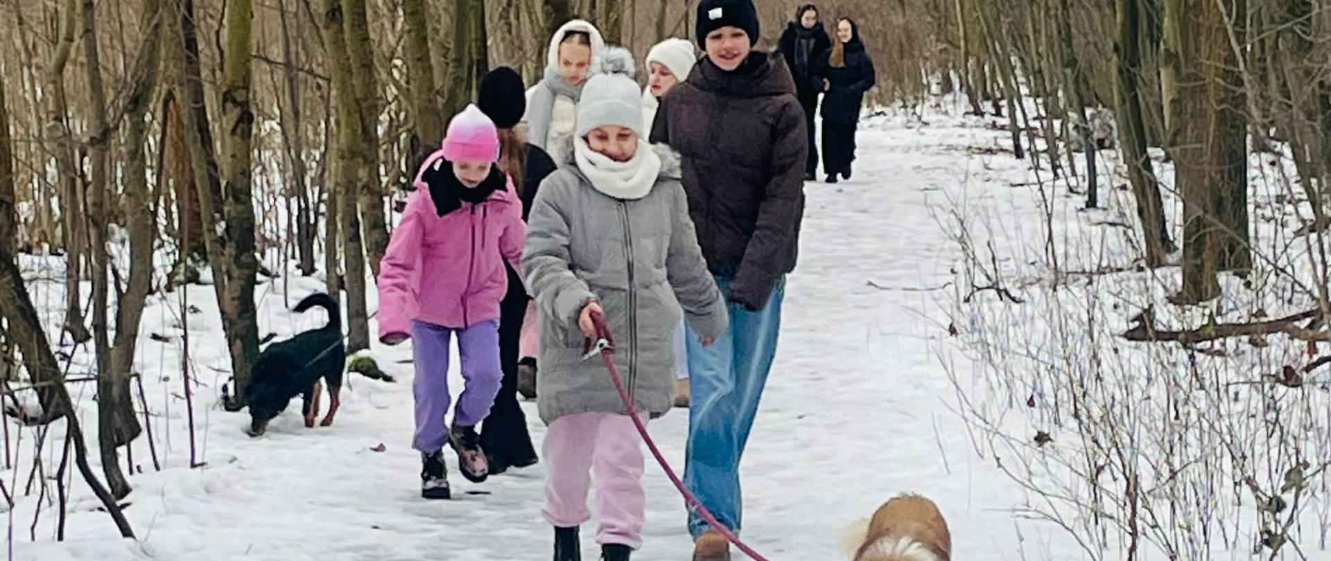 Zdjęcie grupy uśmiechniętych dziewcząt podczas spaceru w lesie z psami fundacji Medor. Dwie z nich trzymają na smyczy psy.
