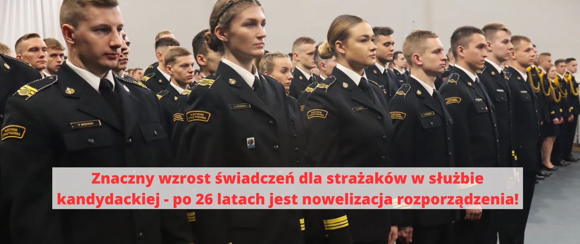 Grupa strażaków w mundurach galowych stoi na sali - na tle zdjęcia napis "Znaczny wzrost świadczeń dla strażaków w służbie kandydackiej - po 26 latach jest nowelizacja rozporządzenia"
