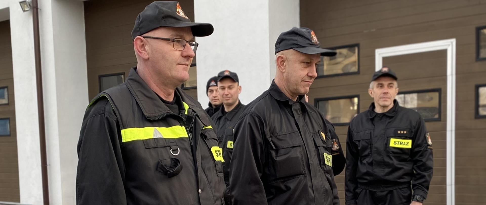 Zdjęcie przestawia przechodzących na emeryturę strażaków. W tle strażacy podziału bojowego jednostki ratowniczo-gaśniczej.