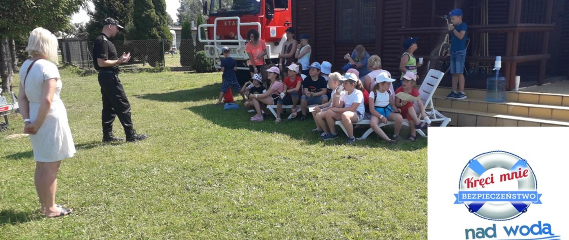 Kolorowe zdjęcie wykonane w jasny dzień, przedstawia na pierwszym planie umundurowanego strażaka w czapce stojącego przodem do grupy siedzących dzieci. obok stoi osoba cywilna. W tle brązowy budynek a obok czerwona kabina ciężarowego pojazdu pożarniczego. W tle drzewa i krzewy. Po prawej stronie wklejone niebieskie logo akcji Kręci mnie bezpieczeństwo nad wodą.
