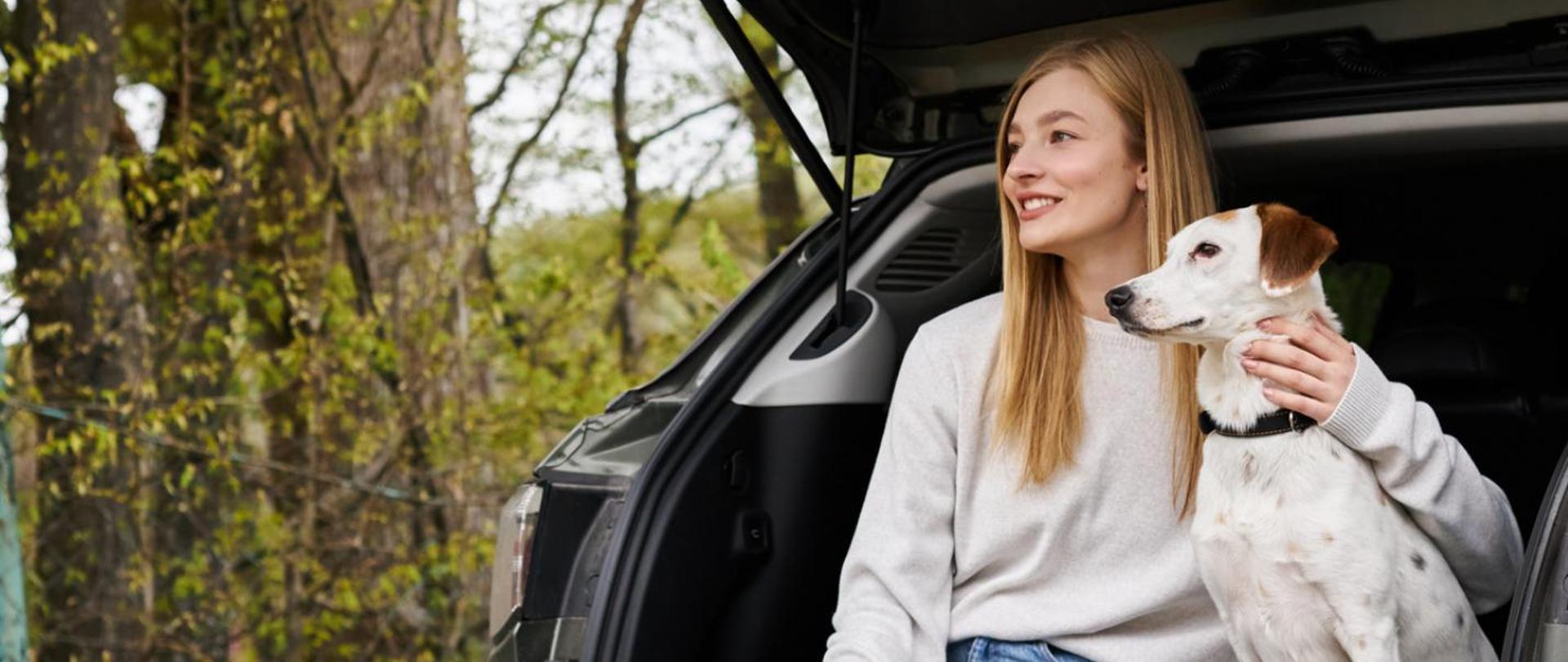 Na zdjęciu jest kobieta i pies, którzy siedzą w bagażniku samochodowym. Znajdują się w lesie.