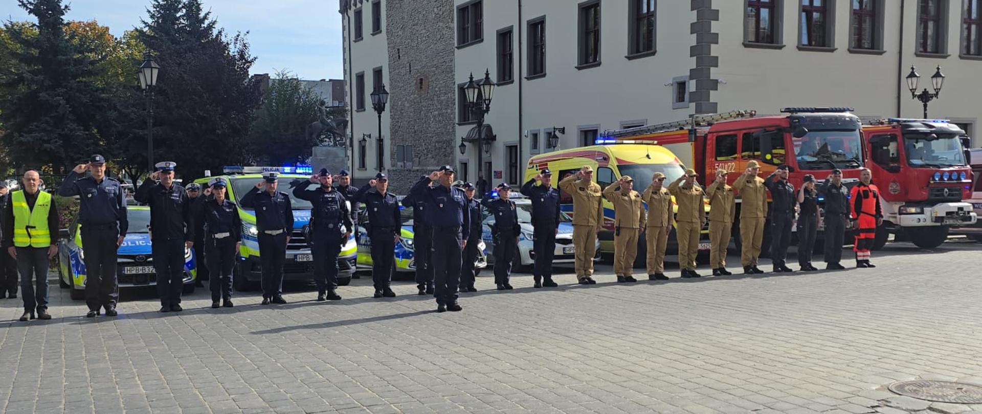 Zdjęcie przedstawia grupę funkcjonariuszy polskich służb ratunkowych i mundurowych, stojących w równym szeregu podczas oficjalnej uroczystości lub apelu na wybrukowanym placu miejskim w tle zabytkowych kamienic.
Po lewej stronie widoczni są funkcjonariusze Policji w ciemnogranatowych mundurach, z których większość salutuje. Za nimi zaparkowane są radiowozy policyjne z włączonymi niebieskimi sygnałami świetlnymi.
W środkowej i prawej części szeregu stoją strażacy z Państwowej Straży Pożarnej w piaskowych (beżowych) mundurach specjalnych oraz czapkach, również salutujący. Obok nich znajduje się ratownik medyczny w jaskrawopomarańczowym kombinezonie. Za strażakami zaparkowany jest żółty ambulans (karetka pogotowia) oraz dwa czerwone wozy strażackie. Po lewej stronie szeregu stoi także mężczyzna w cywilnym ubraniu i odblaskowej, żółtej kamizelce. Wszystkie służby są ustawione przodem do obiektywu, a wydarzenie odbywa się w pełnym świetle dziennym.