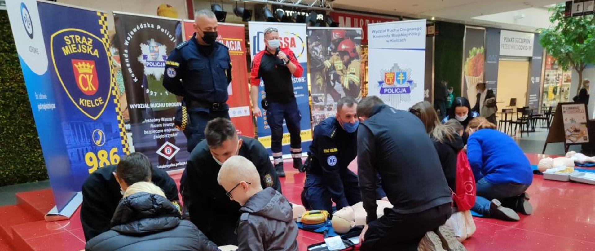 Zdjęcie przedstawia strażaków którzy prezentują jak prawidłowo wykonać masaż serca. Przed nimi znajdują się zainteresowani uczestnicy spotkania.