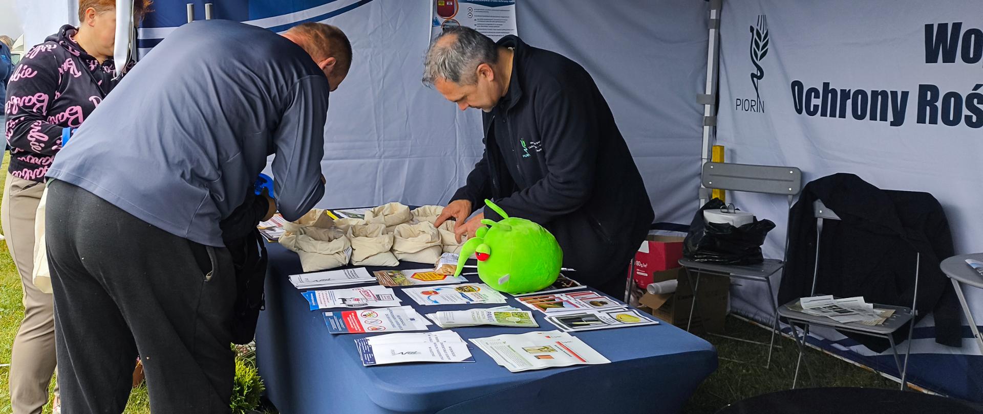 na stole w woreczkach są nasiona, po obu stronach stoją osoby pochylone nad nimi, przed nimi leżą ulotki i zielona maskotka