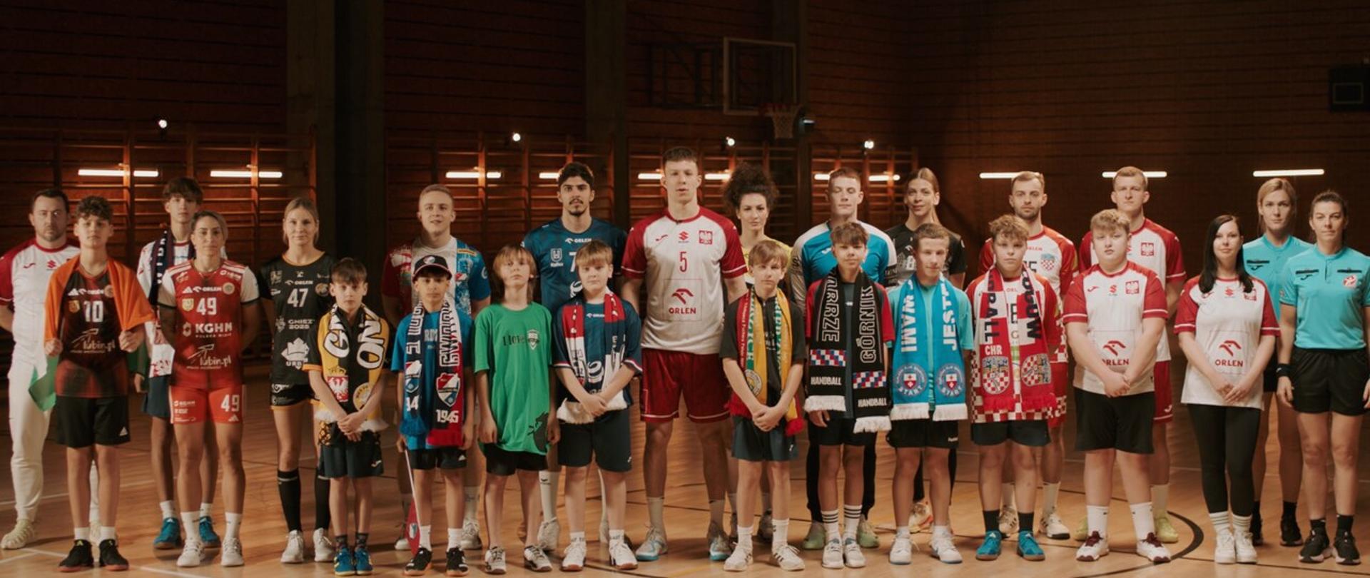 zdjęcie piłkarzy ręcznych z dziećmi na boisku. Grupa ludzi stojąca w kolorowych strojach i szalikach na parkiecie. Z tyłu widać kosze i drabinki
