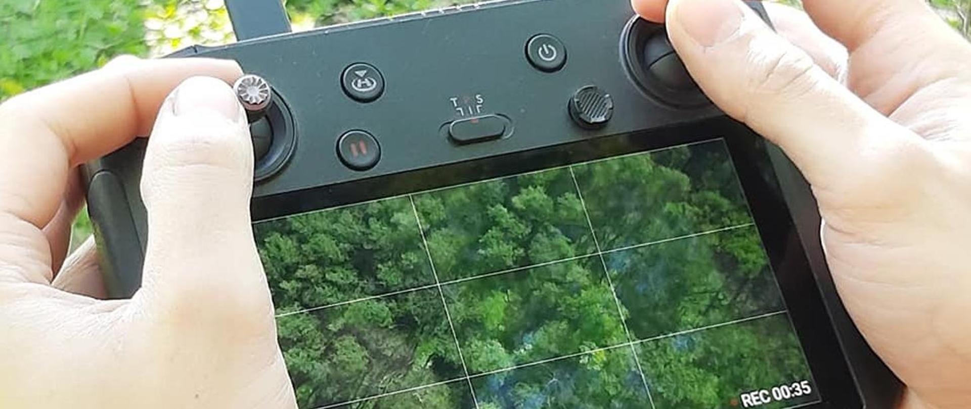 Dłonie trzymające w prostokątne narzędzie z kamerą do obsługi drona. Urządzenie jest czarne, zawiera pokrętła i przyciski. Ma monitor, na którym widać kadr lasu z wysokości i funkcję nagrywania REC. Tłem jest fragment lasu liściastego. Dominuje zielona kolorystyka.