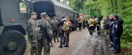 Żołnierze WOT strażacy PSP i OSP i policjanci stoją w grupach w tle pojazdy wojska i straży.