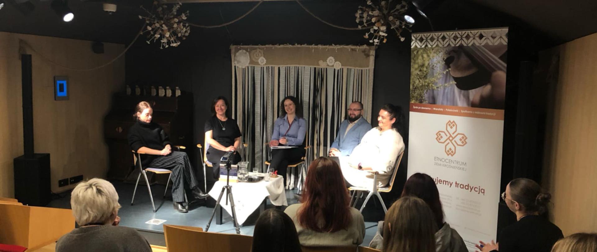 Zdjęcie przedstawia ludzi siedzących na scenie podczas, a naprzeciwko siedzi publiczność. W tle na czarnej ścianie wiszą koronkowe wstążki i wyszywanki.