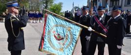 Mężczyzna w wyjściowym mundurze PSP salutuje przed pochulonym sztandarem, który trzyma mężczyzna w wyjściowym mundurze OSP, stojący w środku pocztu sztandarowego, w tle widoczni pozostali uczestnicy uroczystości