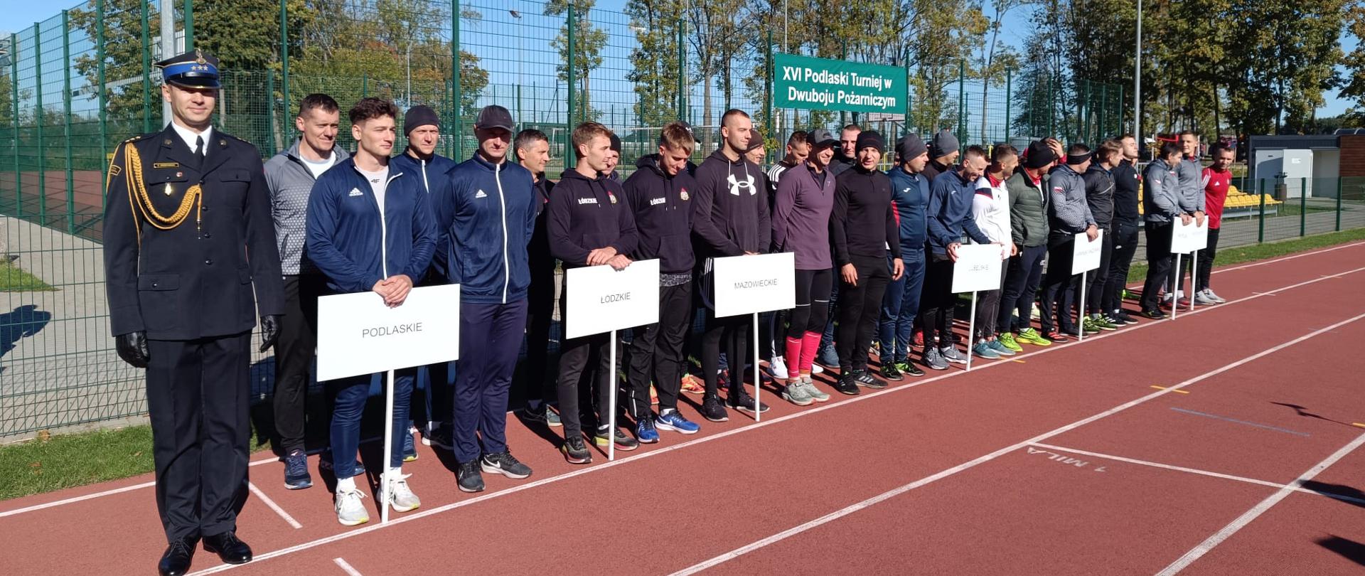 Za nami XVI Podlaski Turniej w Dwuboju Pożarniczym - sportowcy stojący w szeregu, ubrani w dresy - stoją na bieżni i trzymają tabliczki z nazwami województw