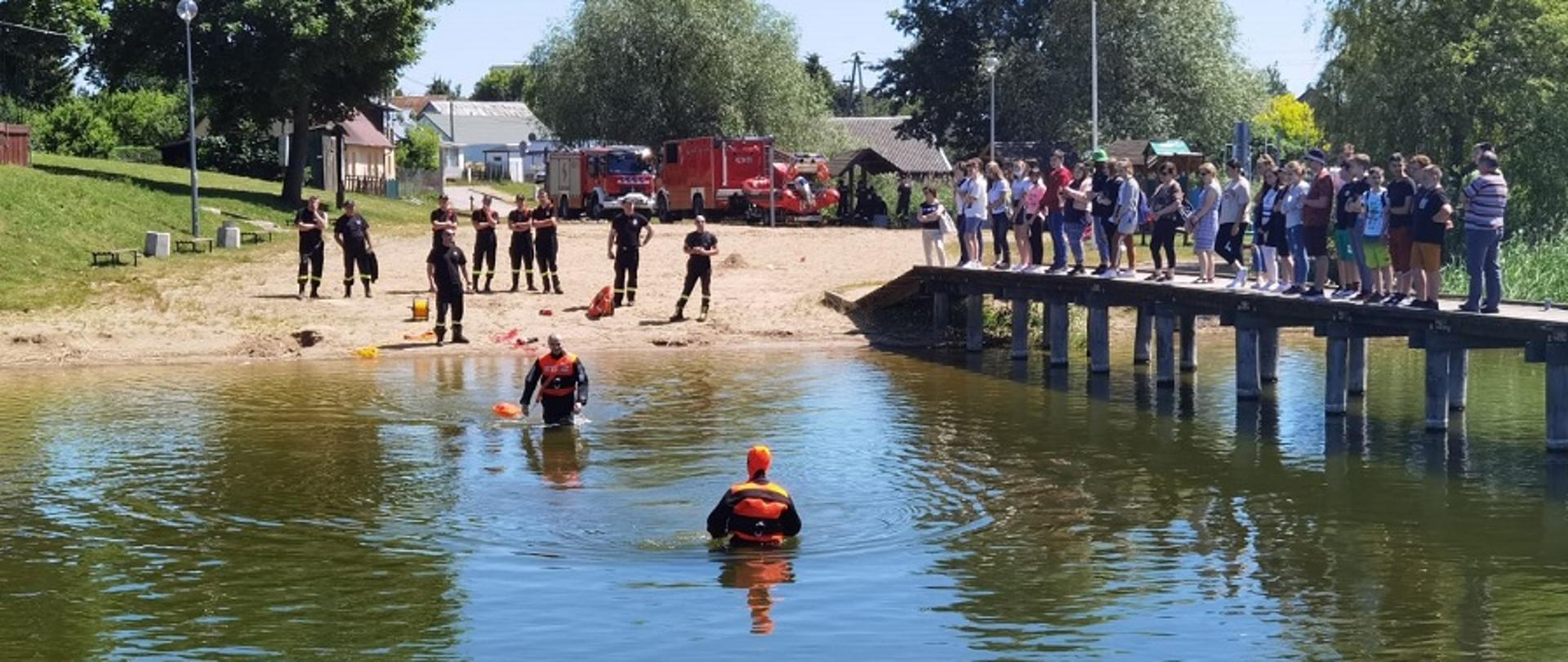 Ludzie stojący na pomoście nad jeziorem. Na brzegu plaży stoją strażacy. W wodzie dwóch strażaków w ubraniach wypornościowych.