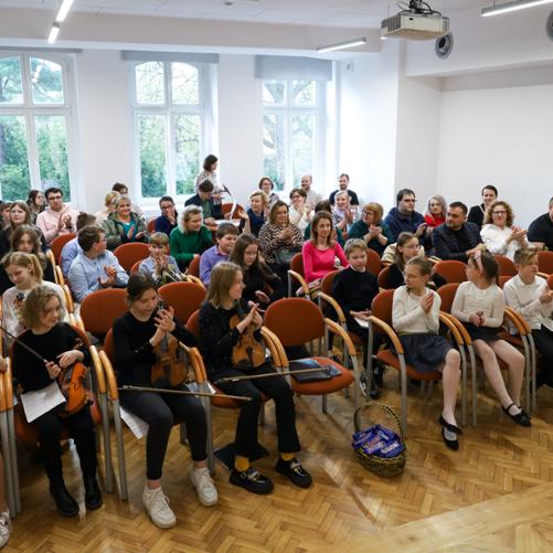 Siedemnasty Szkolny Konkurs Zespołów Kameralnych. Publiczność i wykonawcy zgromadzeni na auli PSM w Głubczycach. Widok ze sceny na publiczność, za oknami widać zazielenione drzewa.