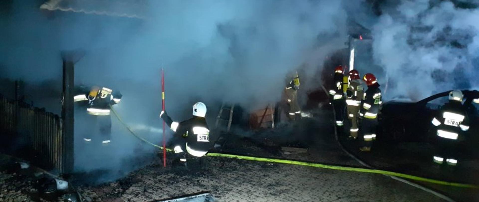 Zdjęcie przedstawia akcję gaszenia pożaru garażu. Na środku grupa strażaków ubranych w ubrania specjalne i hełmy oraz aparaty ODO. Rozwinięte linie gaśnicze. PO prawej stronie stoi spalony samochód. Miejsce jest bardzo gęsto zadymione, teren akcji jest oświetlany przez najaśnicę.