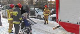 Zdjęcie przedstawia strażaków PSP wiozących starszą kobietę na wózku inwalidzkim w drodze do samochodu wiozacego do punktu szczepień
