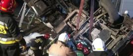 Na zdjęciu widzimy podwozie wywróconego ciągnika siodłowego. Ciągnik znajduje się w rowie, widzimy koła i podwozie. Widzimy sześciu strażaków którzy w pozycji pochylonej próbują dostać się do środka ciągnika. Do podwozia zamocowany jest pas ratowniczy. 