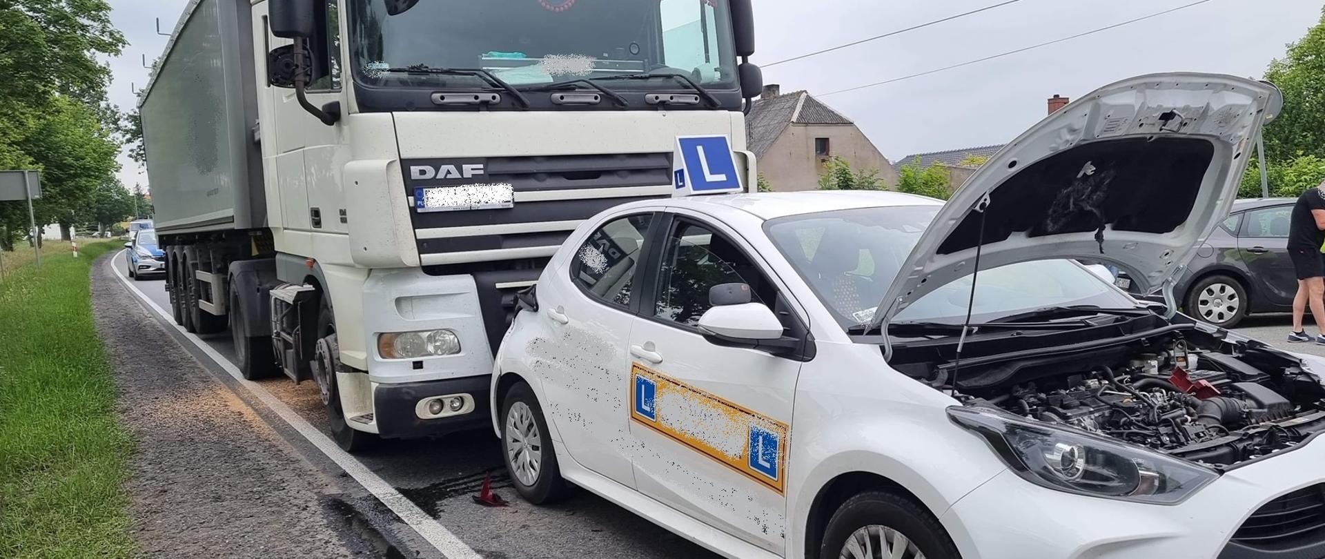 Z prawej strony samochód osobowy koloru białego "nauka jazdy" po kolizji z samochodem ciężarowym koloru białego (s.ciężarowy uderzył w tył s. osobowego). Za samochodem ciężarowym stoi radiowóz policji. 