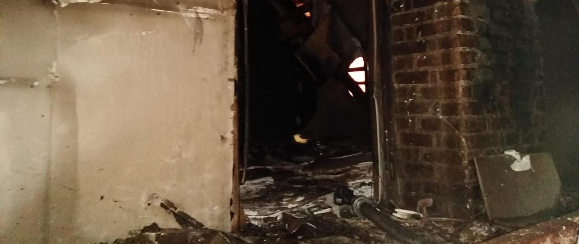 Widok na spalone poddasze przez otwór drzwiowy. Z lewej strony otworu okopcona ściana w kolorze białym, z prawej strony przeówd kominowy wykonany z cegły. W tle drzwi częściowo spalone pokrycie dachu. W drzwiach na ziemi leży odcinek W-52 zakończony prądownicą typu Turbo Jet