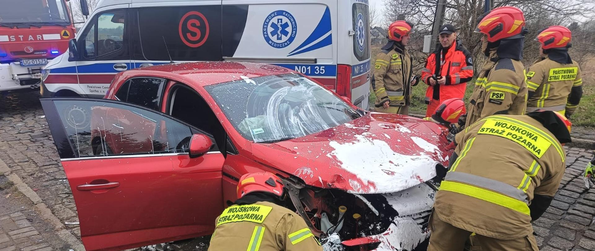 Na zdjęciu czerwony samochód osobowy z uszkodzonym przodem, przy nim strażacy. Dalej karetka i samochód strażacki.