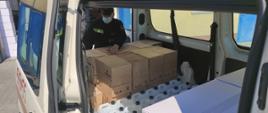 Samochód specjalny – mikrobus, w części ładunkowej pojazdu załadowane kartony z maseczkami, bańki z płynem do dezynfekcji oraz strażak, który dokonuje przeliczenia zabranych środków ochronnych