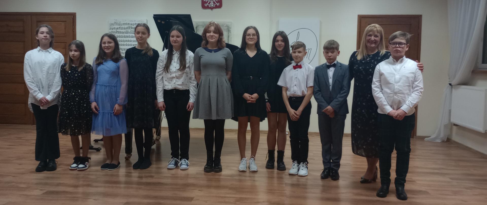 Zdjęcie zbiorowe przedstawiające uczestników koncertu klasy fortepianu dodatkowego. Na brązowej drewnianej scenie stoją uczniowie i uczennice których jest 11, z prawej strony wśród nich stoi ich nauczycielka. W tle widać klapę fortepianu oraz wiszące godło państwowe.