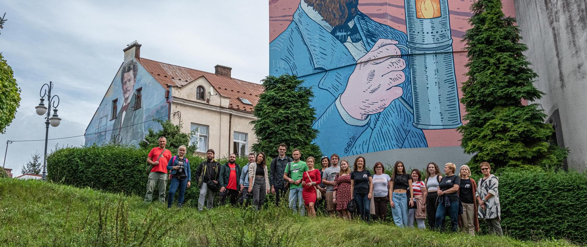 Słuchacze i nauczyciele stojący na tle ścian budynków na których są namalowane postacie Ignacego Łukasiewicza i Jana Szczepanika. 