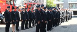 Strażacy ochotniczych straży pożarnych stoją obok siebie w dwóch rzędach za nimi ustawione są samochody pożarnicze.