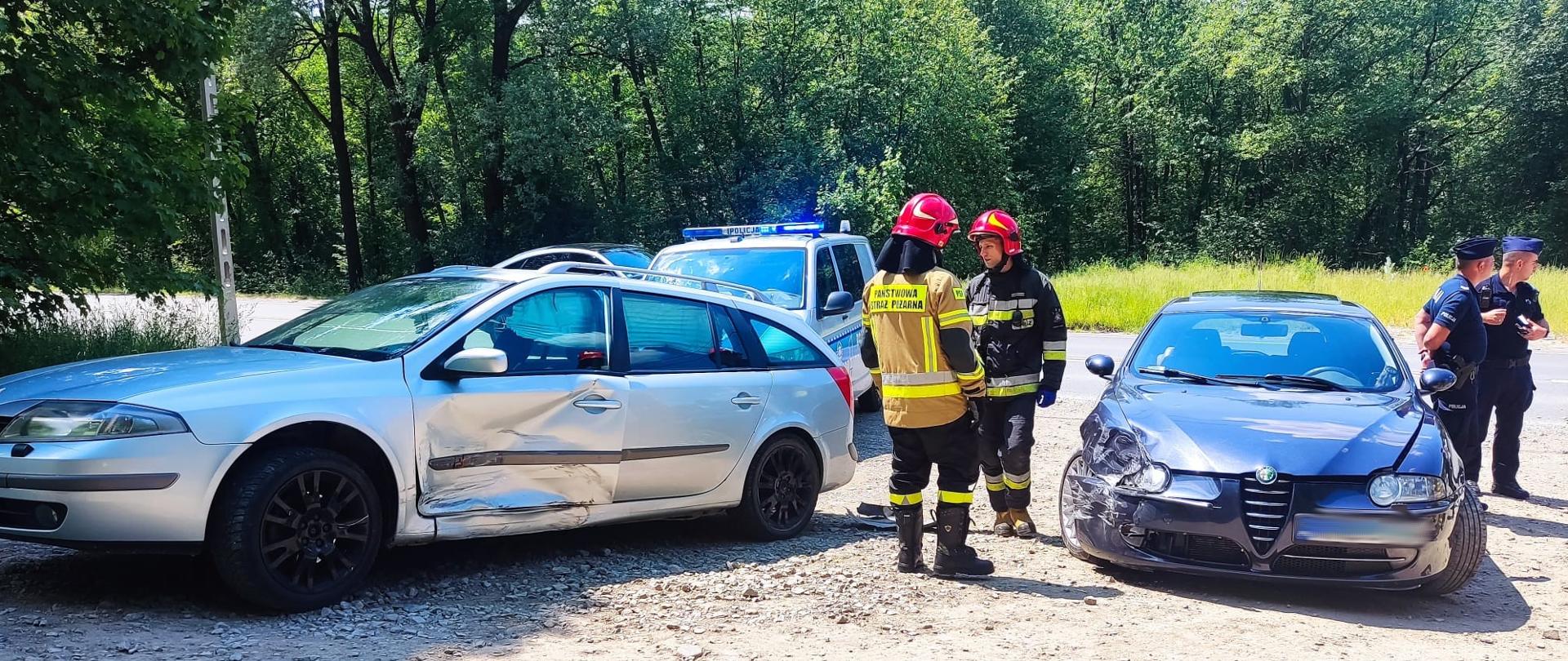 Dwa uszkodzone samochody na poboczu a obok nich strażacy i policjanci