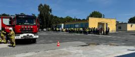 Z lewej strony zdjęcia stoi pojazd pożarniczy. W prawej części stoją wszyscy pracownicy zakładu, którzy zostali ewakuowani z budynku.