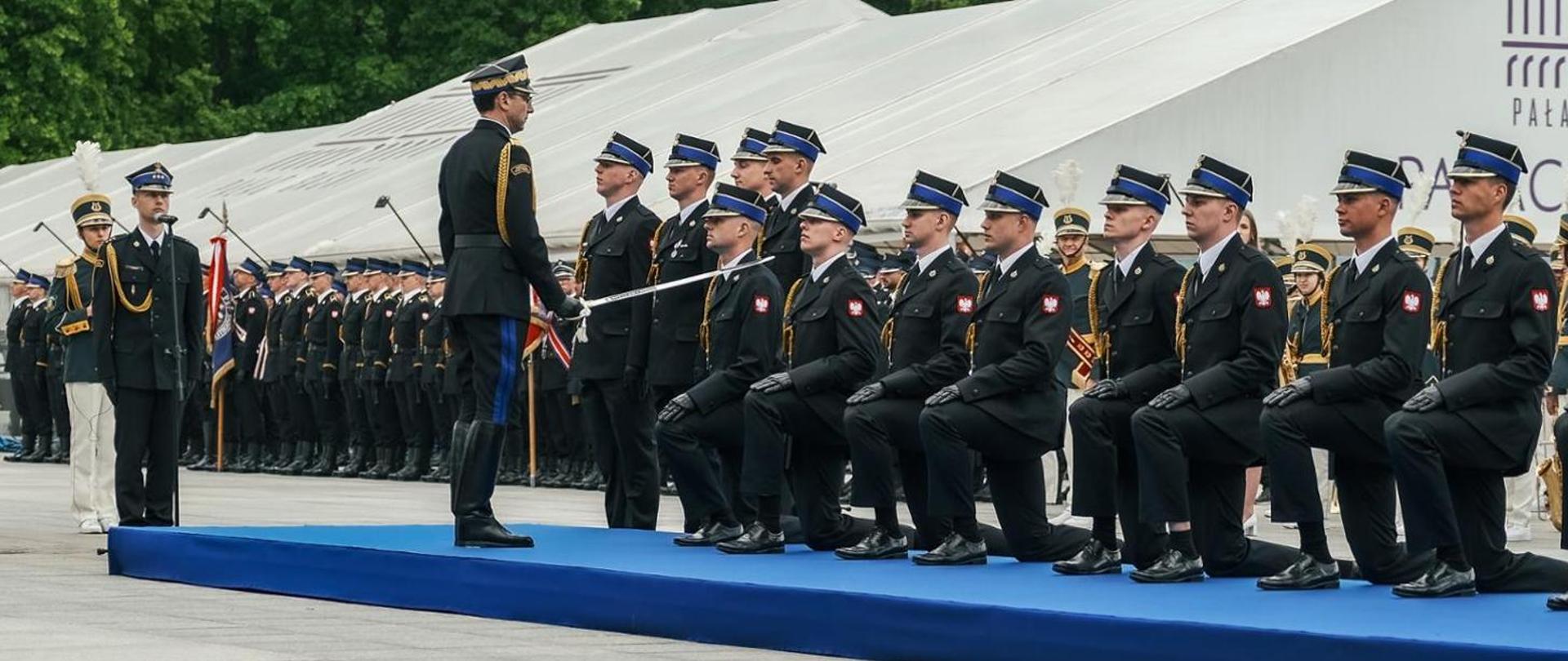 Z lewej Komendant Główny z szablą w dłoni, z prawej klękający na podeście rząd awansowanych strażaków. Wszyscy w mundurach wyjściowych. 