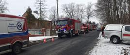 Samochody gaśnicze straży pożarnej zaparkowane na drodze przy posesji na której doszło do pożaru. Miejsce zdarzenia ogrodzone czerwonymi pachołkami. Widać także karetkę pogotowia ratunkowego.